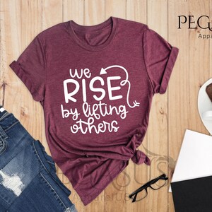 Pu&ograve; includere: Una maglietta di colore marrone rossiccio con il testo "We Rise by lifting others" in lettere bianche. La maglietta &egrave; piegata e mostrata su una superficie di legno.