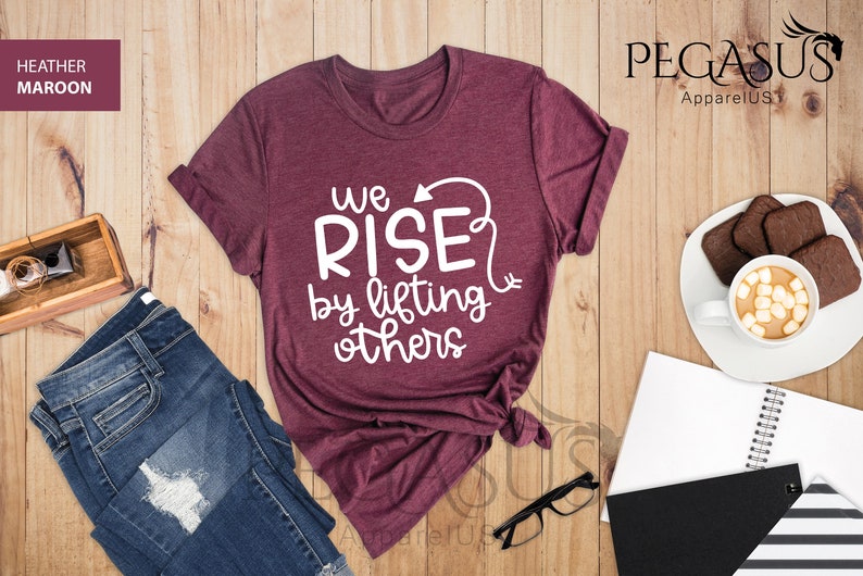 Pu&ograve; includere: Una maglietta di colore marrone rossiccio con il testo "We Rise by lifting others" in lettere bianche. La maglietta &egrave; piegata e mostrata su una superficie di legno.