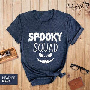 May include: Heather navy blue t-shirt with the words "SPOOKY SQUAD" in white, along with a jack-o'-lantern face design. The shirt is displayed on a wooden surface, with a small wooden box, glasses, and a wallet in the background.