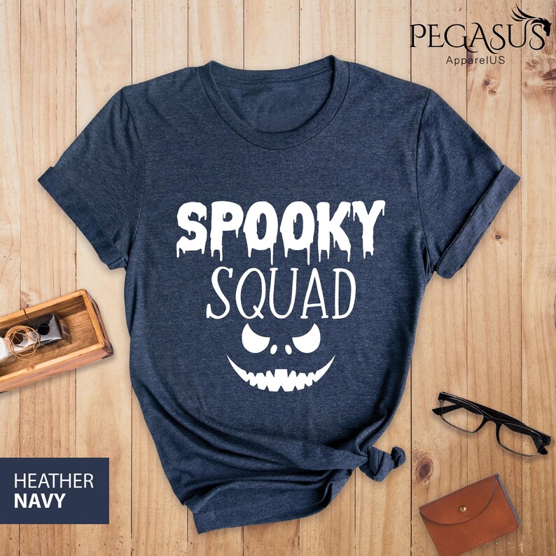 May include: Heather navy blue t-shirt with the words "SPOOKY SQUAD" in white, along with a jack-o'-lantern face design. The shirt is displayed on a wooden surface, with a small wooden box, glasses, and a wallet in the background.