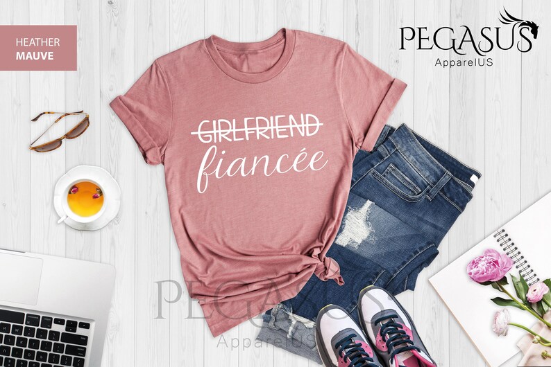 May include: A heather mauve t-shirt with the text "GIRLFRIEND" crossed out and "fianc&eacute;e" written below it. The shirt is laid out on a white wooden surface with a pair of jeans, a pair of sneakers, a cup of tea, sunglasses, and a notebook with flowers.