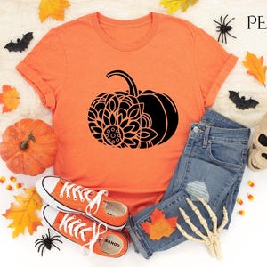 May include: A heather orange t-shirt featuring a black mandala pumpkin design. The shirt is laid out on a white surface with jeans, a skull, and autumn leaves.