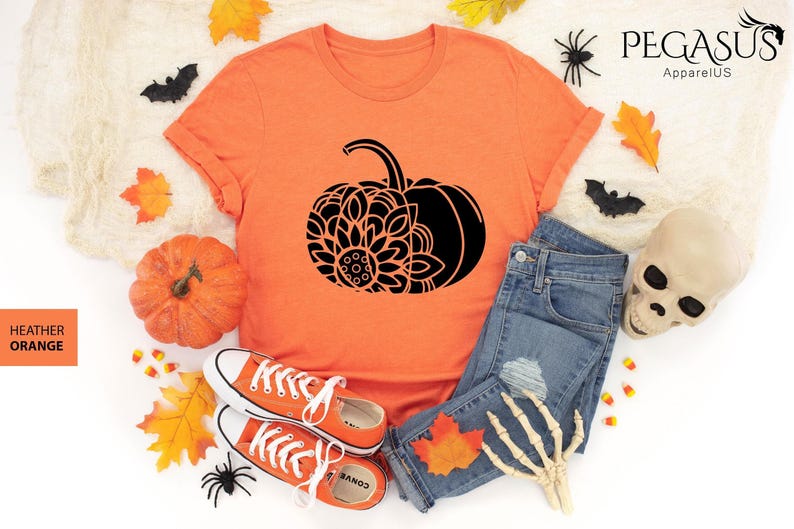 May include: A heather orange t-shirt featuring a black mandala pumpkin design. The shirt is laid out on a white surface with jeans, a skull, and autumn leaves.