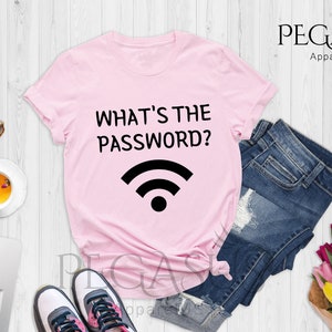 May include: Pink t-shirt with the text "What's the password?" and a wifi symbol. The shirt is laid out on a white wooden surface with a pair of sunglasses, a cup of tea, a laptop, a pair of sneakers, a pair of denim shorts, and a bouquet of flowers.