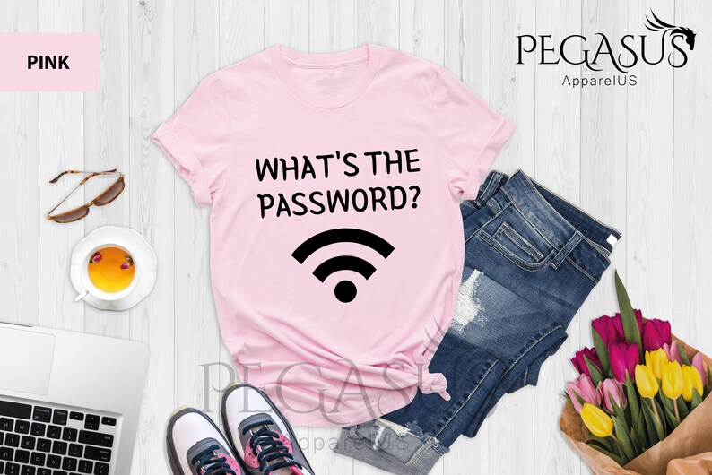 May include: Pink t-shirt with the text "What's the password?" and a wifi symbol. The shirt is laid out on a white wooden surface with a pair of sunglasses, a cup of tea, a laptop, a pair of sneakers, a pair of denim shorts, and a bouquet of flowers.