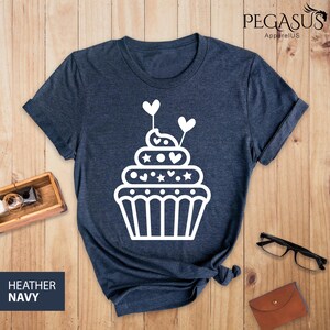 May include: A heather navy blue t-shirt with a white cupcake graphic. The cupcake has white hearts and stars on it.