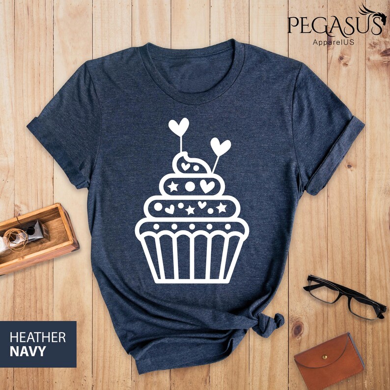 May include: A heather navy blue t-shirt with a white cupcake graphic. The cupcake has white hearts and stars on it.