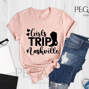 May include: A heather peach t-shirt with the text "Girls Trip Nashville" in black. The design includes hearts and a cowboy boot. The shirt is paired with blue jeans and sneakers.