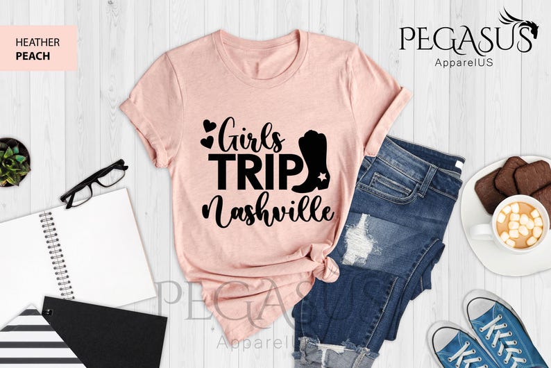 May include: A heather peach t-shirt with the text "Girls Trip Nashville" in black. The design includes hearts and a cowboy boot. The shirt is paired with blue jeans and sneakers.