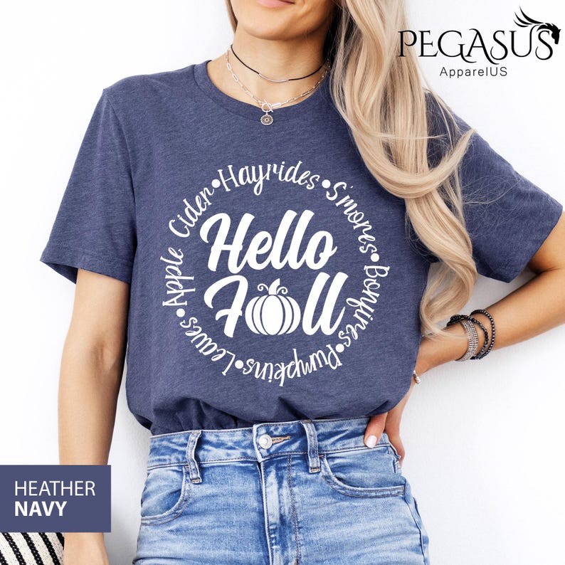 May include: Heather navy blue t-shirt with a white circular design that says "Hello Fall" with a pumpkin in the center. The design is surrounded by words like "Hayrides" and "Pumpkins". The shirt is made of a soft material.