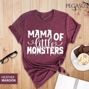 May include: Heather maroon t-shirt with the text "MAMA OF little MONSTERS" in white. The shirt is styled with rolled-up sleeves and a knotted hem. The words "Heather Maroon" are printed on a small rectangular label.