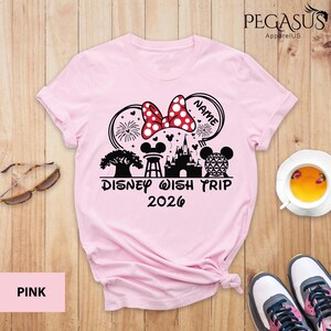 May include: Pink t-shirt with a Disney-themed graphic. The design includes a Minnie Mouse bow, fireworks, a castle, and the text "Disney Wish Trip 2026". The word "NAME" is in a speech bubble. Sunglasses, a cup of tea, and sneakers are also visible.