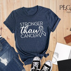 May include: A heather navy blue t-shirt with the text "Stronger than Cancer" and a white ribbon graphic. The shirt is laid flat on a wooden surface with a pair of jeans, a notebook, and a pair of glasses.