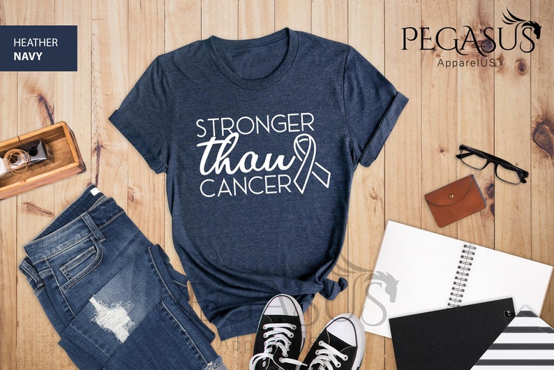 May include: A heather navy blue t-shirt with the text "Stronger than Cancer" and a white ribbon graphic. The shirt is laid flat on a wooden surface with a pair of jeans, a notebook, and a pair of glasses.