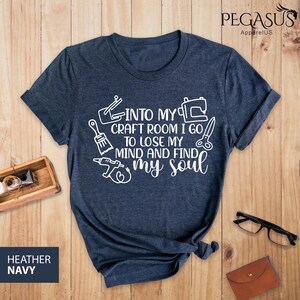 K&ouml;nnte beinhalten: Ein heidefarbenes marineblaues T-Shirt mit wei&szlig;em Text, der "Into my craft room I go to lose my mind and find my soul" lautet. Der Text ist von Bildern von Bastelwerkzeugen umgeben, darunter ein Pinsel, eine N&auml;hmaschine, eine Schere und eine Klebepistole.