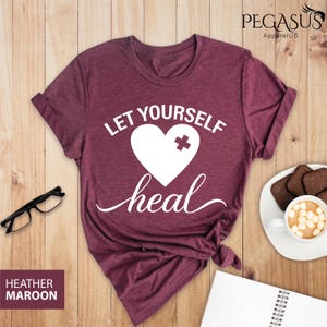 May include: Heather maroon t-shirt with white text that reads "Let Yourself Heal" and a white heart with a white plus sign over it.