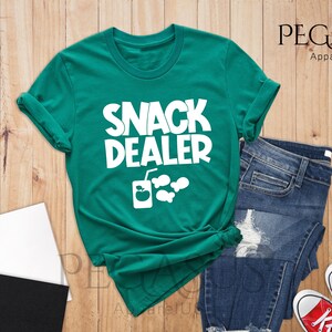 Peut inclure: Un t-shirt vert avec le texte "Snack Dealer" en blanc. Le t-shirt a un graphique d'une boisson et de poissons.