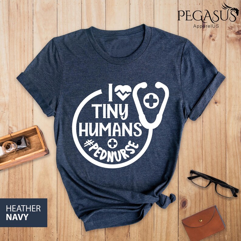 Op de afbeelding: Een heideblauw t-shirt met een wit grafisch ontwerp. De grafiek toont een stethoscoop met een hart en de tekst "Ik hou van kleine mensen #pednurse".