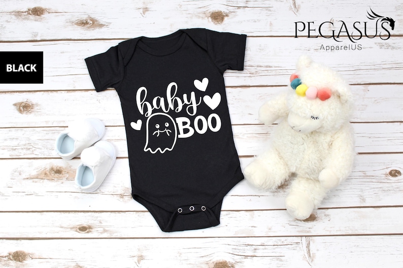 May include: Black baby bodysuit with the words "baby BOO" and a ghost graphic in white. The bodysuit is next to a pair of white baby shoes with blue interiors and a white stuffed animal with a rainbow headband.