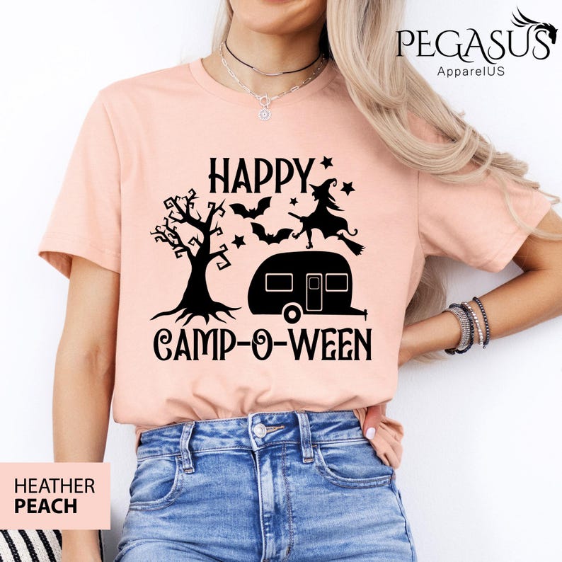 May include: Peach-coloured t-shirt with a black graphic that reads "HAPPY CAMP-O-WEEN." The design includes a tree, bats, a witch on a broom, and a camper. The shirt is made of a soft material and is perfect for Halloween.
