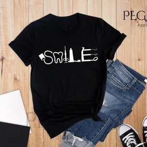 May include: Black t-shirt with a white graphic of dental tools spelling out the word "SMILE".