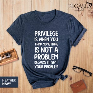 May include: A dark blue t-shirt with the text "Privilege is when you think something is not a problem because it isn't your problem" printed in white.