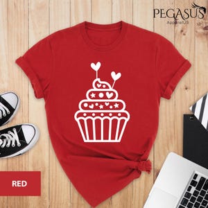 May include: Red t-shirt with a white cupcake design. The cupcake has white frosting with hearts and stars on it. The cupcake is topped with a white cherry.