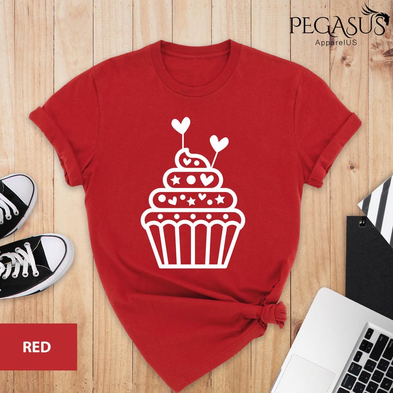 May include: Red t-shirt with a white cupcake design. The cupcake has white frosting with hearts and stars on it. The cupcake is topped with a white cherry.