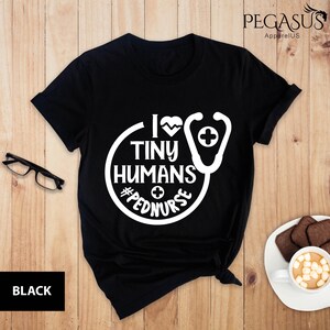Op de afbeelding: Zwart T-shirt met een witte cirkelvormige afbeelding met de tekst "I love tiny humans #pednurse" met een stethoscoop en een hartvormige afbeelding.