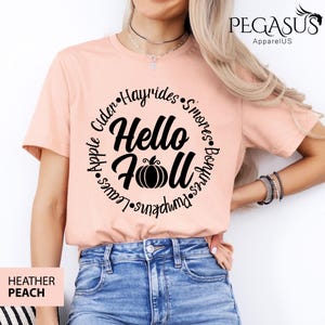 May include: Heather peach t-shirt with a circular design that says "Hello Fall" with a pumpkin graphic. The design is surrounded by words like "Apple", "Hayrides", and "Pumpkins". The shirt is a short-sleeved crew neck.
