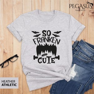 Pu&ograve; includere: T-shirt grigio atletico heather con grafica nera che dice "So Franken Cute" con pipistrelli e un design mostruoso di Frankenstein. La maglietta &egrave; abbinata a jeans blu e occhiali.