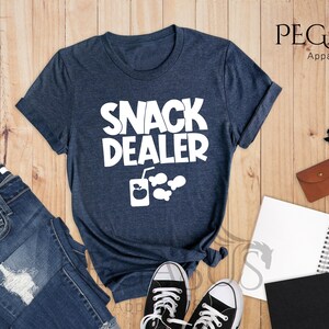 Peut inclure: Un t-shirt bleu marine chin&eacute; avec le texte "Snack Dealer" en blanc. Le t-shirt pr&eacute;sente &eacute;galement un graphique blanc d'une bo&icirc;te de jus avec une paille et trois poissons.