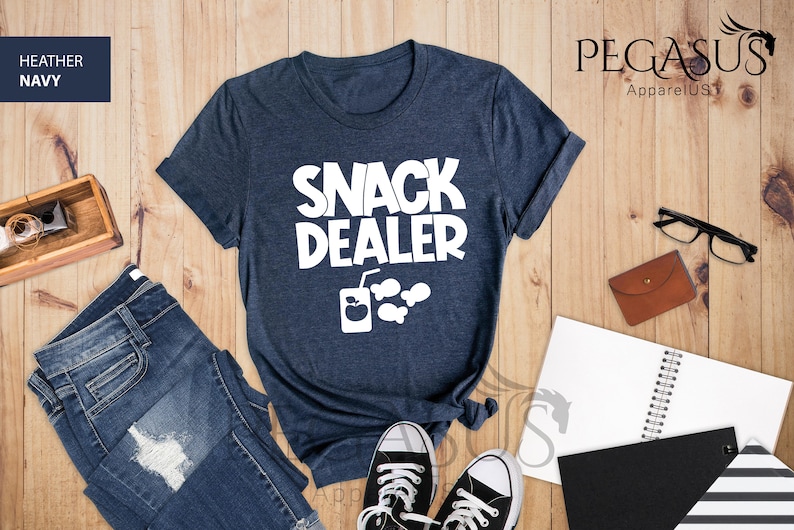 Peut inclure: Un t-shirt bleu marine chin&eacute; avec le texte "Snack Dealer" en blanc. Le t-shirt pr&eacute;sente &eacute;galement un graphique blanc d'une bo&icirc;te de jus avec une paille et trois poissons.
