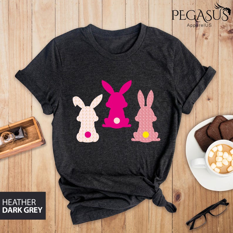 May include: Dark heather grey t-shirt featuring three bunny rabbit designs in pink and white. The bunnies have patterned bodies and white or yellow tail accents. The shirt is from Pegasus ApparelUS.
