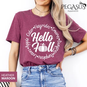 May include: Heather maroon t-shirt with a white circular design that says "Hello Fall" with a pumpkin graphic. The design is surrounded by words like "Hayrides" and "Pumpkins". The shirt is paired with blue jeans.