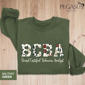 May include: Military green sweatshirt with the letters "BCBA" in a floral design. Below the letters is the text "Board Certified Behavior Analyst". The sweatshirt has a crew neck and long sleeves.