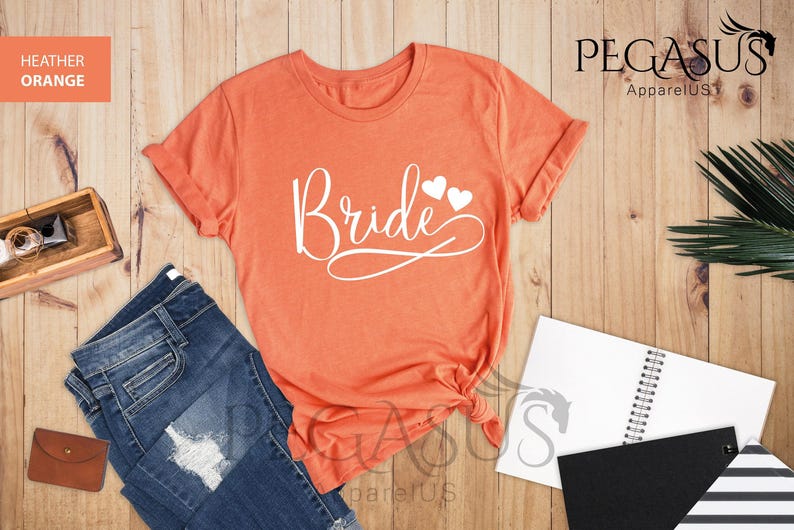 May include: A heather orange t-shirt with the word "Bride" in white script with two small white hearts above the word. The t-shirt is on a wooden surface with a pair of jeans, a notebook, and a plant.