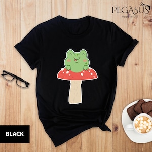 May include: Black t-shirt with a green frog sitting on a red and white mushroom. The frog is smiling and has its tongue sticking out.