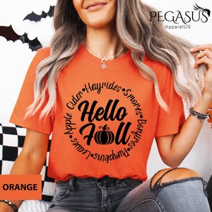 May include: Orange t-shirt with the text "Hello Fall" in a circular design, including a pumpkin graphic. The design also features words like "Hayrides" and "Pumpkins". The shirt is a short-sleeved crew neck.