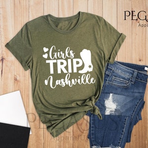 May include: Heather olive green t-shirt with the text "Girls Trip Nashville" in white. The shirt is styled with blue jeans, a notebook, and a cup of coffee with marshmallows. The shirt has a rolled sleeve.