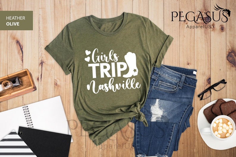 May include: Heather olive green t-shirt with the text "Girls Trip Nashville" in white. The shirt is styled with blue jeans, a notebook, and a cup of coffee with marshmallows. The shirt has a rolled sleeve.