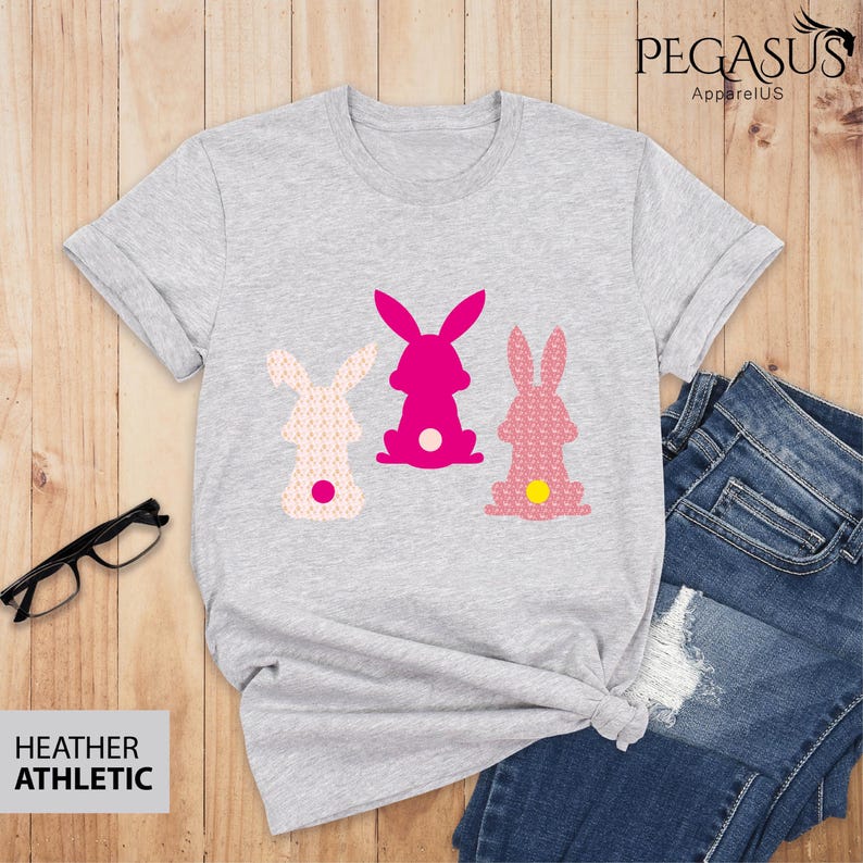 May include: Heather gray t-shirt featuring three bunny silhouettes in pink and patterned pink. The shirt has rolled sleeves and is paired with blue jeans. The text "HEATHER ATHLETIC" is visible.