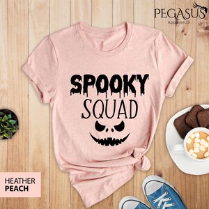 May include: Heather peach-colored t-shirt with the words "Spooky Squad" in black, featuring a jack-o'-lantern face. The shirt is folded at the bottom. The image also shows a pair of blue sneakers, a plate with cookies, and a cup of hot chocolate.