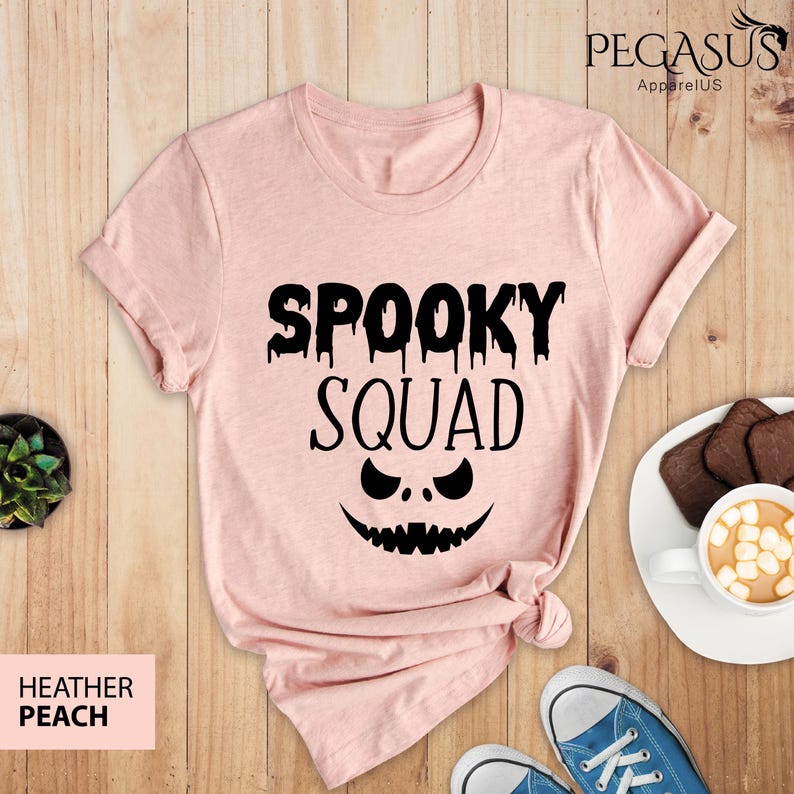 May include: Heather peach-colored t-shirt with the words "Spooky Squad" in black, featuring a jack-o'-lantern face. The shirt is folded at the bottom. The image also shows a pair of blue sneakers, a plate with cookies, and a cup of hot chocolate.