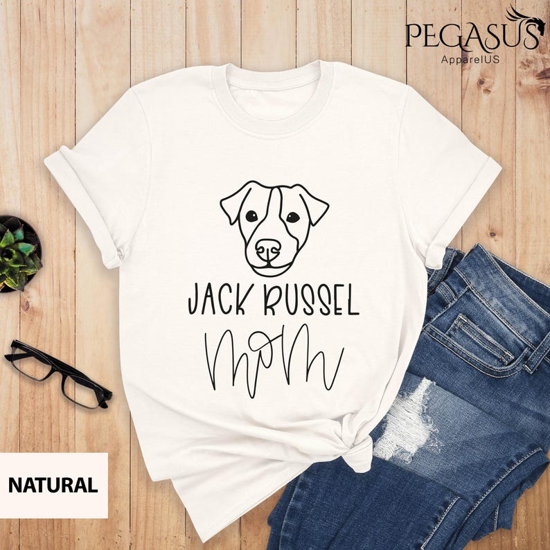 May include: White t-shirt with a black line drawing of a Jack Russell Terrier's face and the words "JACK RUSSEL MOM" in black script. The shirt is paired with blue jeans. The brand name "PEGASUS ApparelUS" is in the upper right corner.