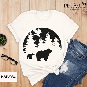Puede incluir: Camiseta blanca con una silueta negra de un oso y un cachorro en una escena de bosque. El dise&ntilde;o est&aacute; en un c&iacute;rculo con un p&aacute;jaro volando sobre los &aacute;rboles. El texto "NATURAL" est&aacute; en la esquina inferior izquierda.