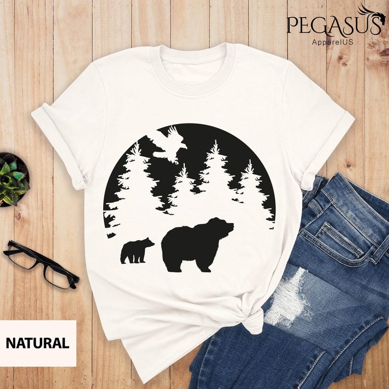 Puede incluir: Camiseta blanca con una silueta negra de un oso y un cachorro en una escena de bosque. El dise&ntilde;o est&aacute; en un c&iacute;rculo con un p&aacute;jaro volando sobre los &aacute;rboles. El texto "NATURAL" est&aacute; en la esquina inferior izquierda.