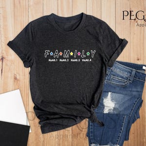 May include: Dark heather grey t-shirt with the word "FAMILY" in white letters with colorful stars between each letter. The text "NAME 1 NAME 2 NAME 3 NAME 4" is printed below the word "FAMILY".