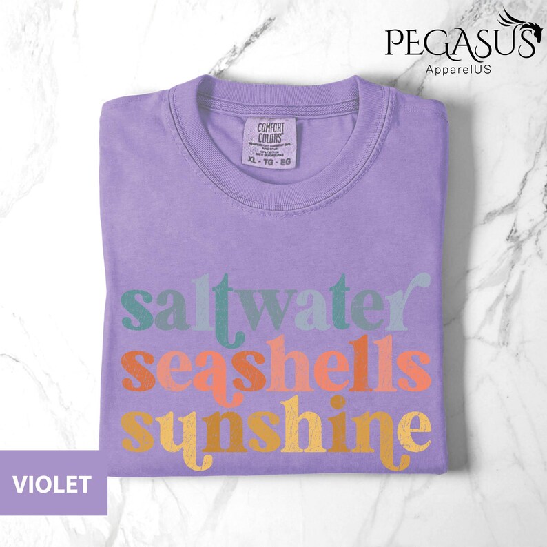 May include: A violet Comfort Colors t-shirt with the words "saltwater seashells sunshine" in a colorful, retro font. The shirt is folded neatly on a marble surface. The tag reads XL-TG-EG.