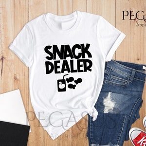 Peut inclure: T-shirt blanc avec un texte noir qui dit "Snack Dealer" et un graphique d'une bo&icirc;te de jus avec une pomme et quelques en-cas.